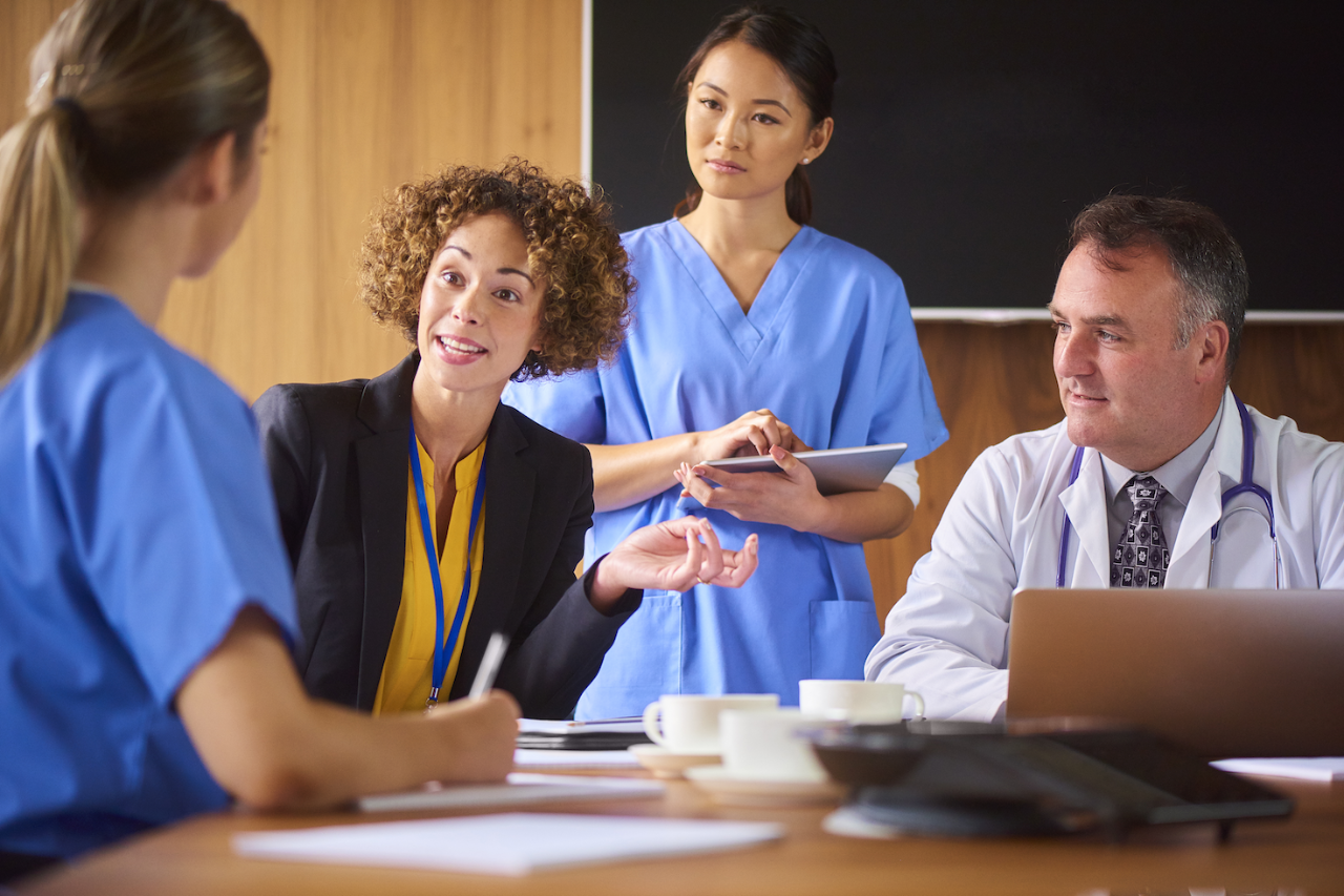 a senior doctor chats to his nursing team around a boardroom table. He is accompanied by  a woman in suits who could be the hospital administrator , salesperson or lawyer .